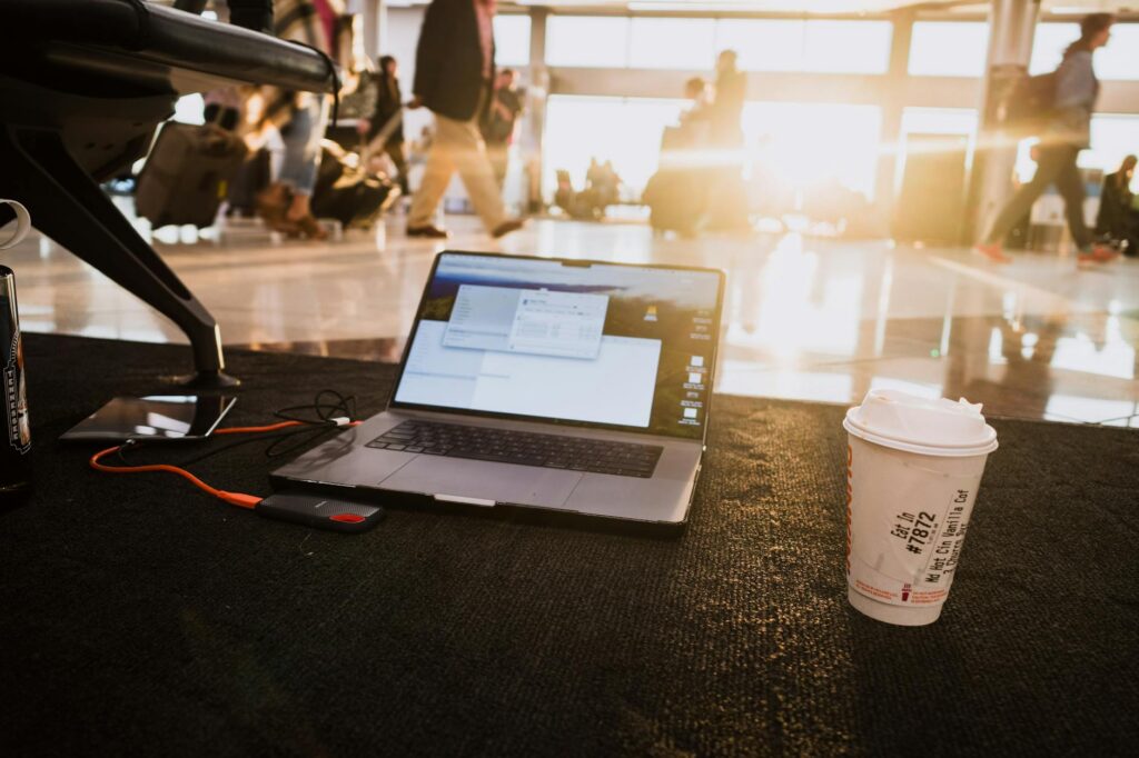 Laptop and coffee cup on airport floor with passengers walking by in warm sunlight.